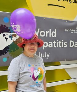 Claire Martin standing in front of a World Hepatitis Day Banner wearing an orange hat and gret t-shirt
